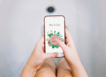 Une femme dans une baignoire tient un smartphone affichant l’application My S Life, partenaire de Saforelle, dédiée à la santé intime. L’écran montre un visuel floral. Le slogan « Plus j’en sais, mieux je me sens » est affiché en bas de l’image en lettres blanches sur fond rose.